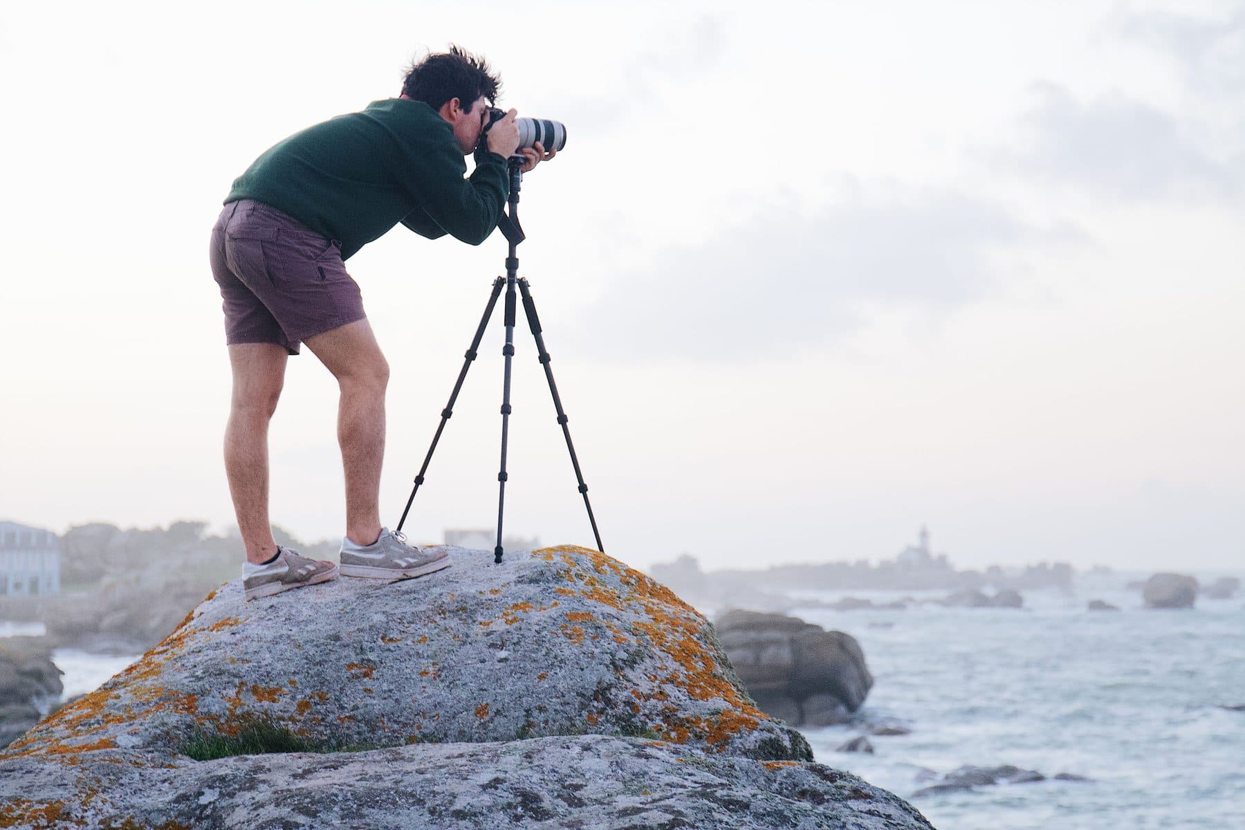 A photographer braces a tripod-mounted camera on a coastal rock at sunrise
